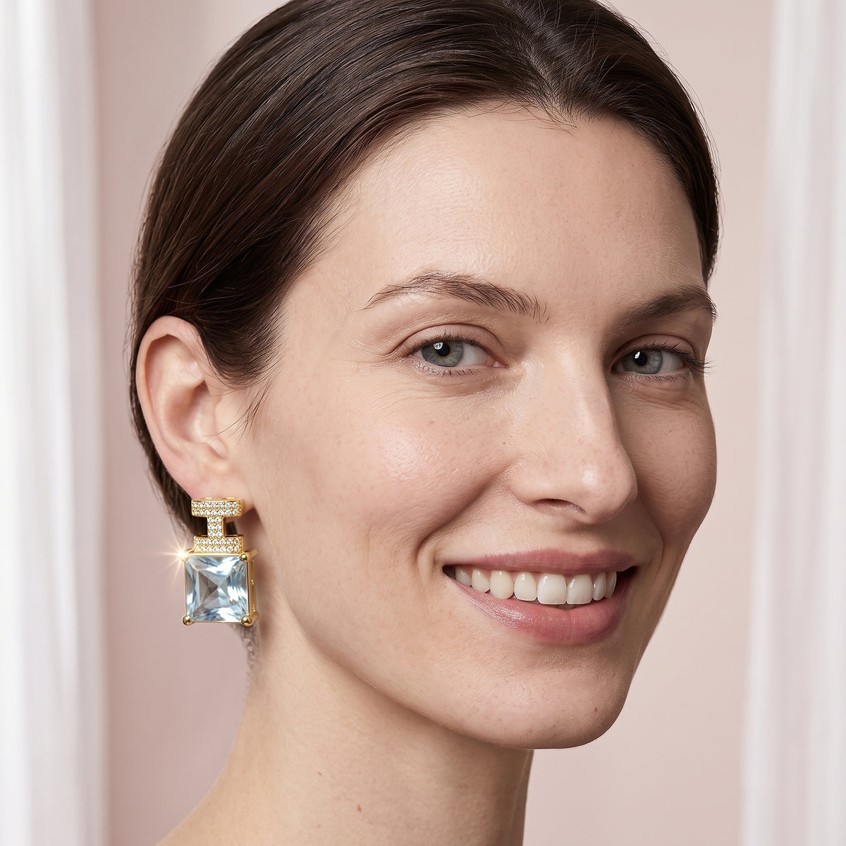 Modelo sorrindo usando o brinco cravejado com pedra azul clara banhado a ouro da ALUENA