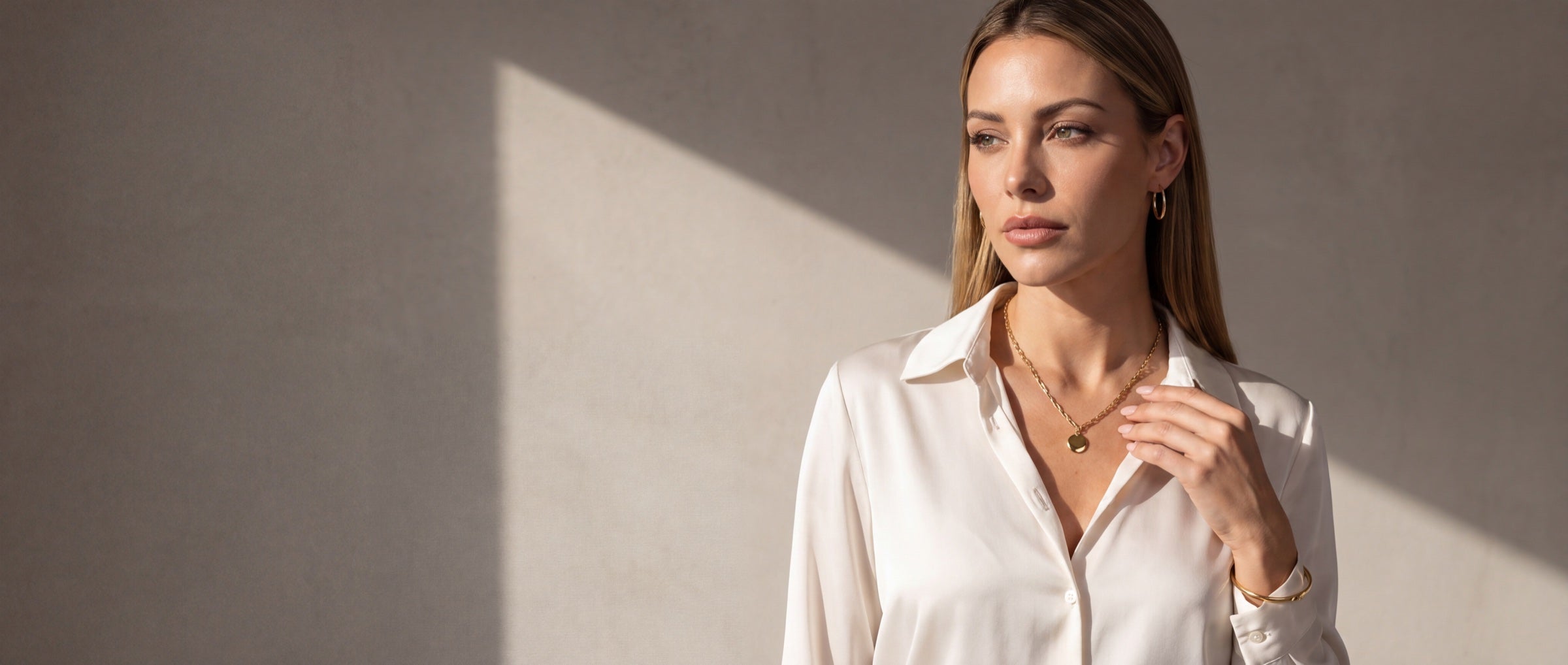 Mulher de blusa de seda marfim, olhar sereno, usando colar com pingente redondo, argolas e bracelete banhados a ouro, diante de uma parede clara com luz de fim de tarde